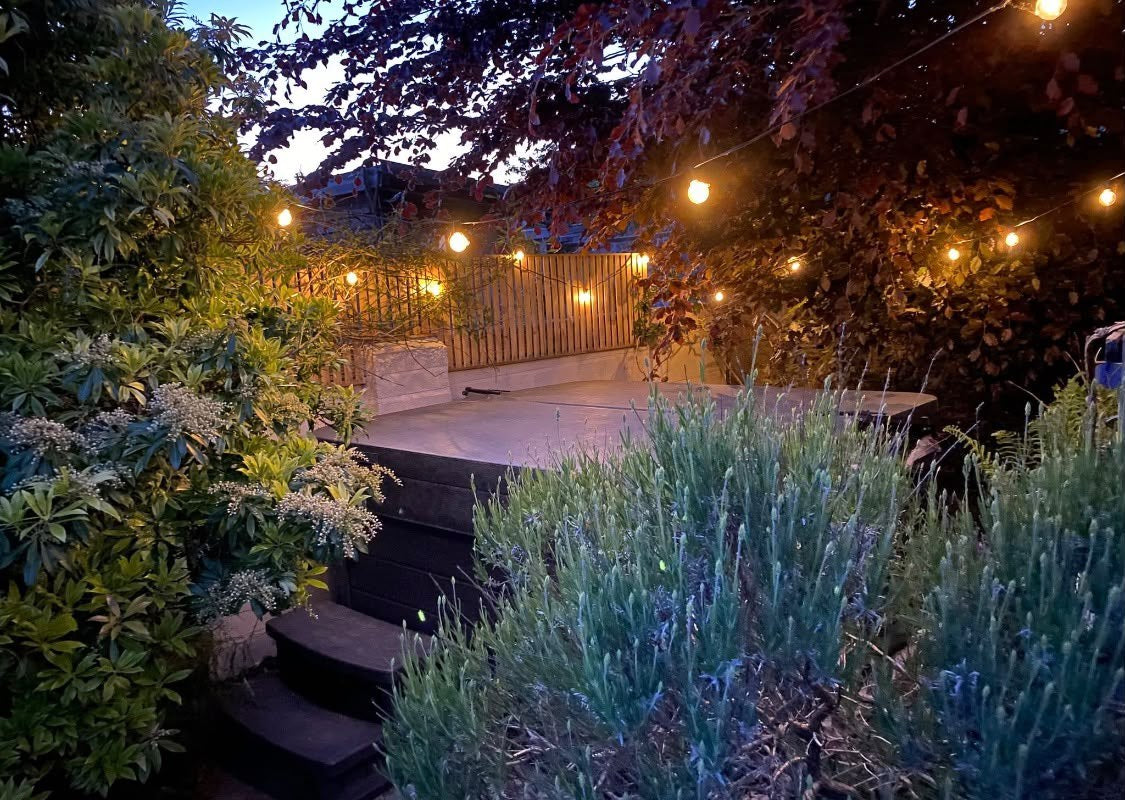 A timber outdoor bar with a pergola roof, pendant lighting and a BBQ, beside a patio with table and chairs and planted raised beds