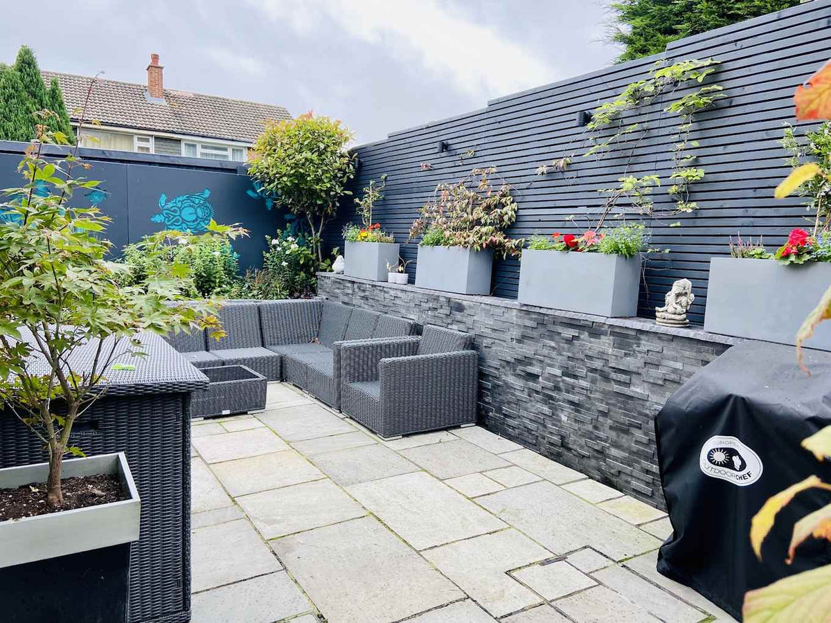 A modern courtyard with sandstone paving, a slate faced wall and planted grey troughs