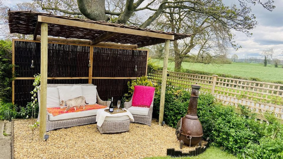 A timber pergola with a woven hazel roof and screen, rattan seating on gravel and a chiminea, with open fields beyond the fence