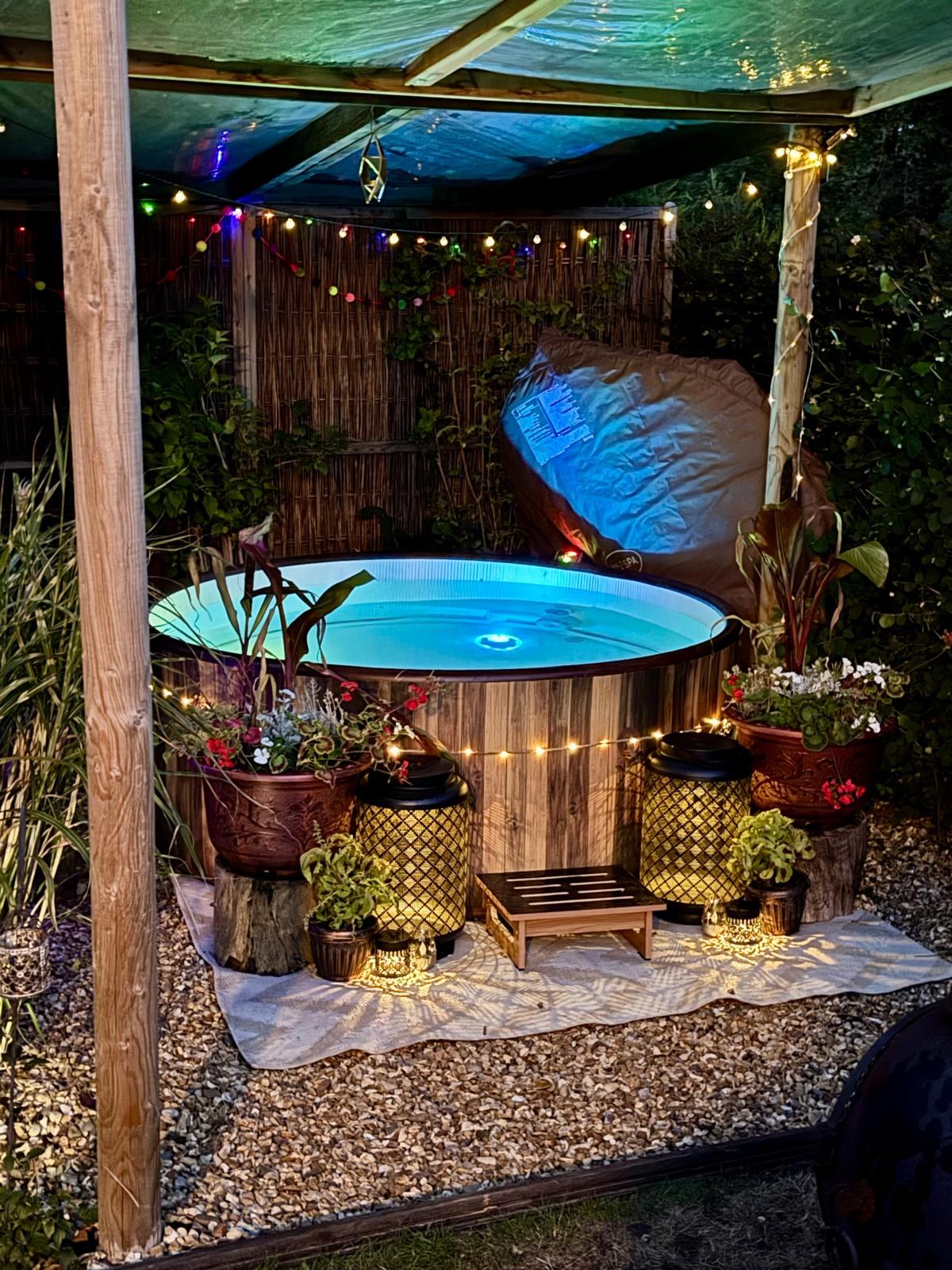 Karen's own wooden barrel hot tub lit up at night under a hazel pergola with lanterns and planted pots