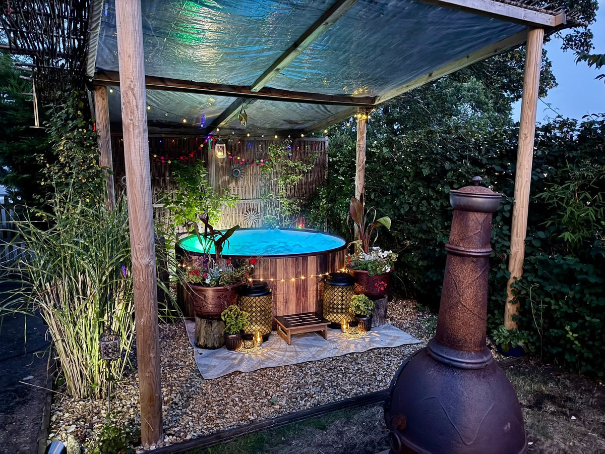 A wooden barrel hot tub lit up at dusk under a timber pergola, with festoon lights, lanterns and planted pots