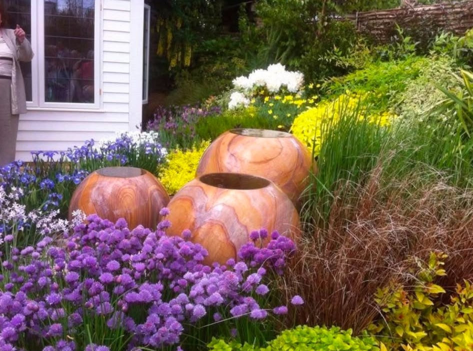 Three sandstone urns set among chives, alliums and grasses beside a white boarded wall